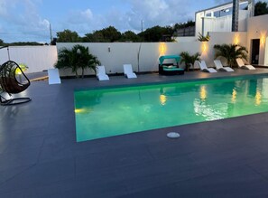 A swimming pool with lounge chairs and a hanging chair, surrounded by a white wall and greenery.