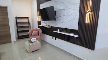 Ein modernes Wohnzimmer mit einem an der Wand mit Marmoroptik befestigten Flachbildfernseher, einem rosafarbenen Sessel, einem Glastisch und einer Holzregaleinheit.