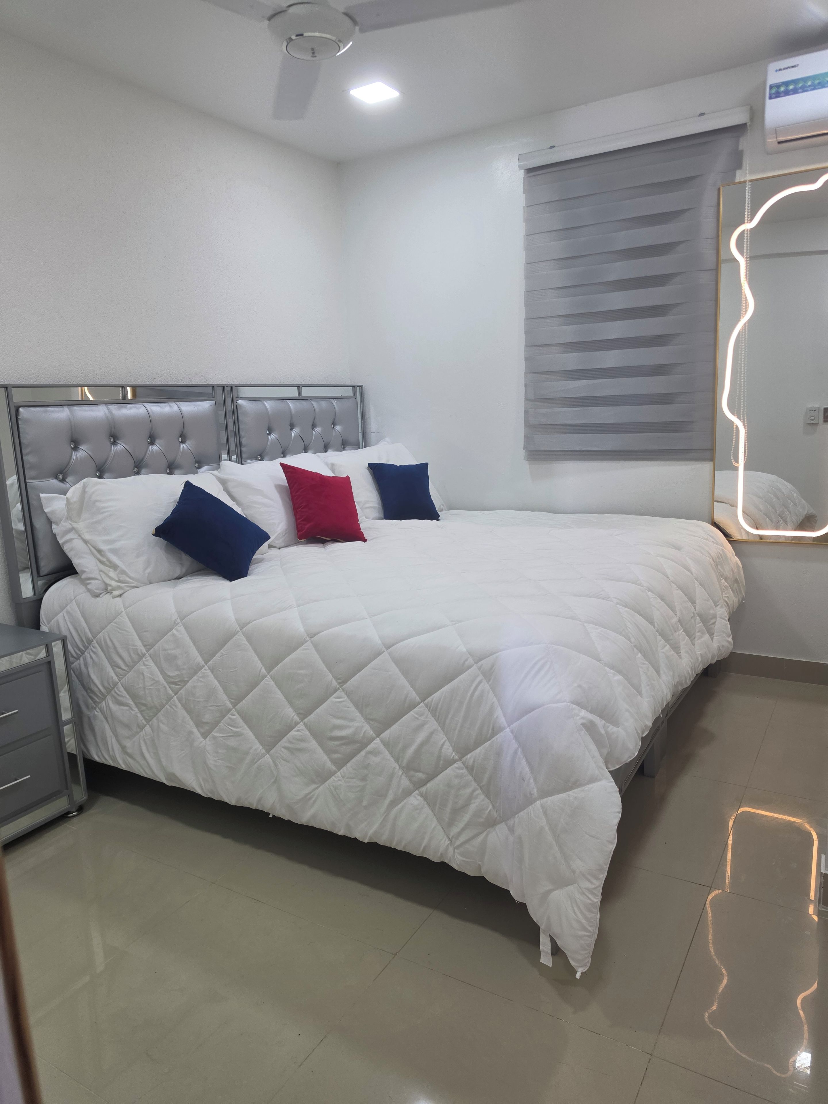 A neatly made bed with a quilted white duvet, a grey upholstered headboard, and a bedside table with a grey cabinet.