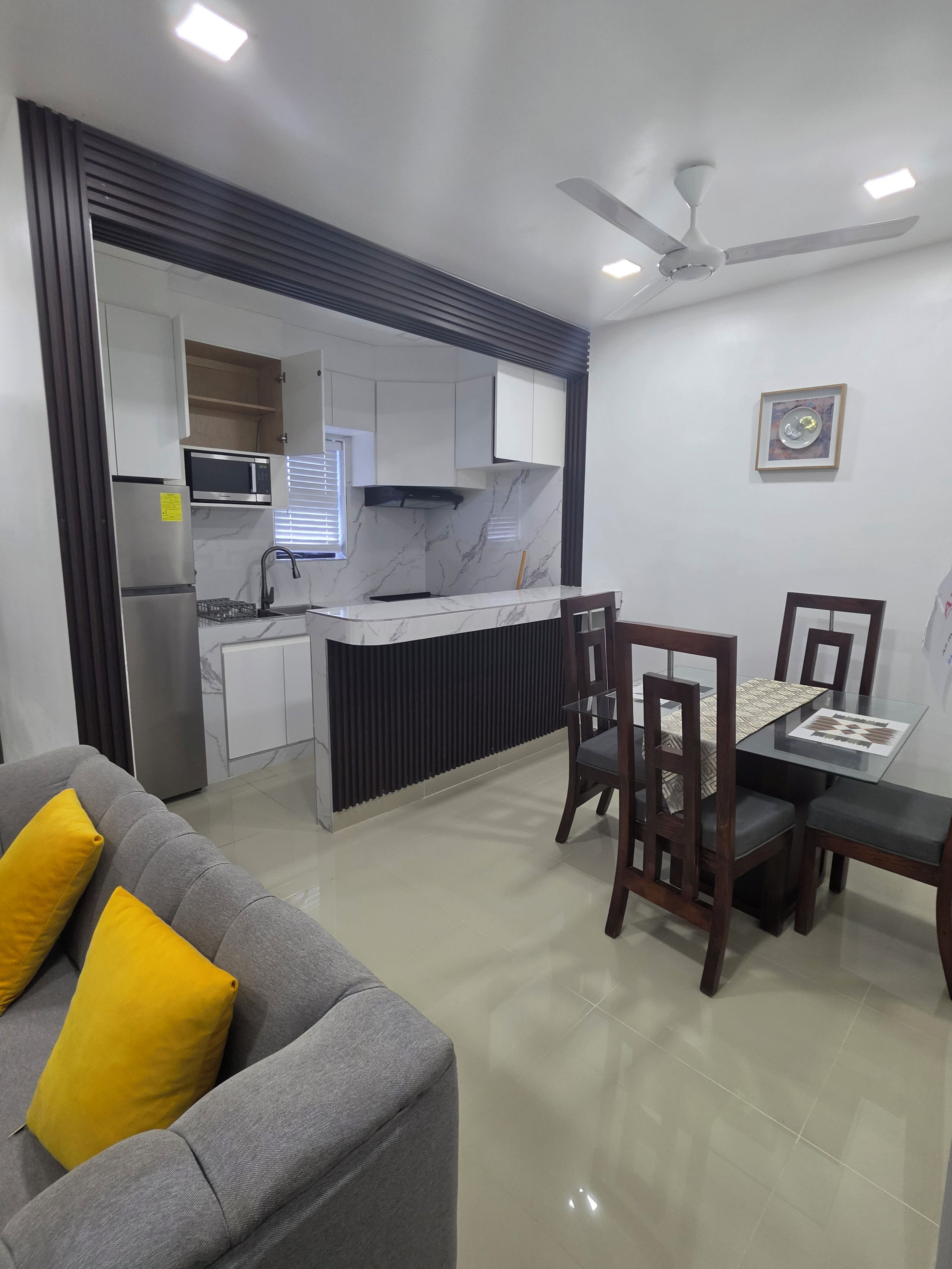 Una cocina moderna con microondas integrado, un comedor con mesa de vidrio y sillas de madera, y un sofá gris con cojines amarillos.