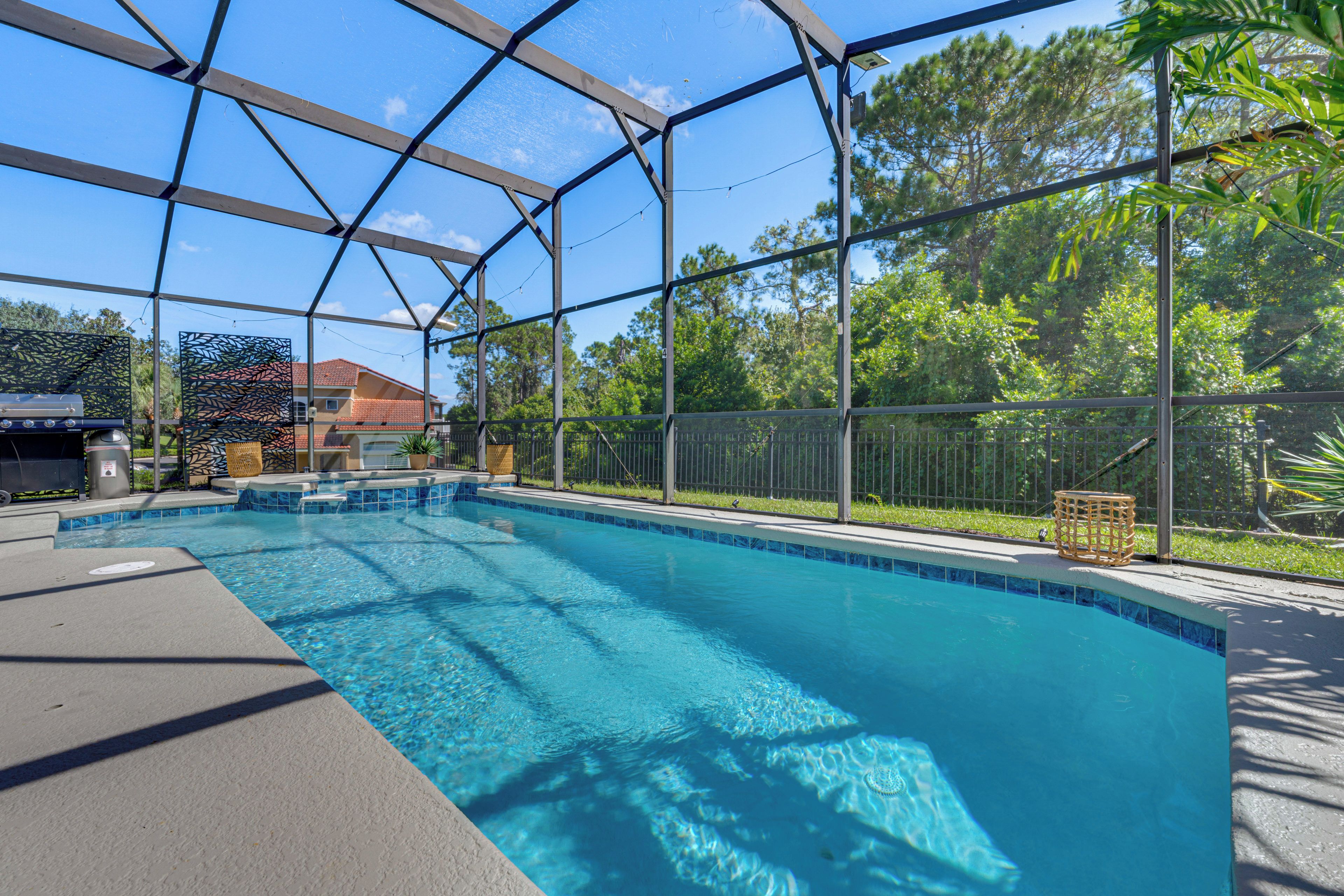 A clear blue pool with a covered area, surrounded by trees and a house in the background.