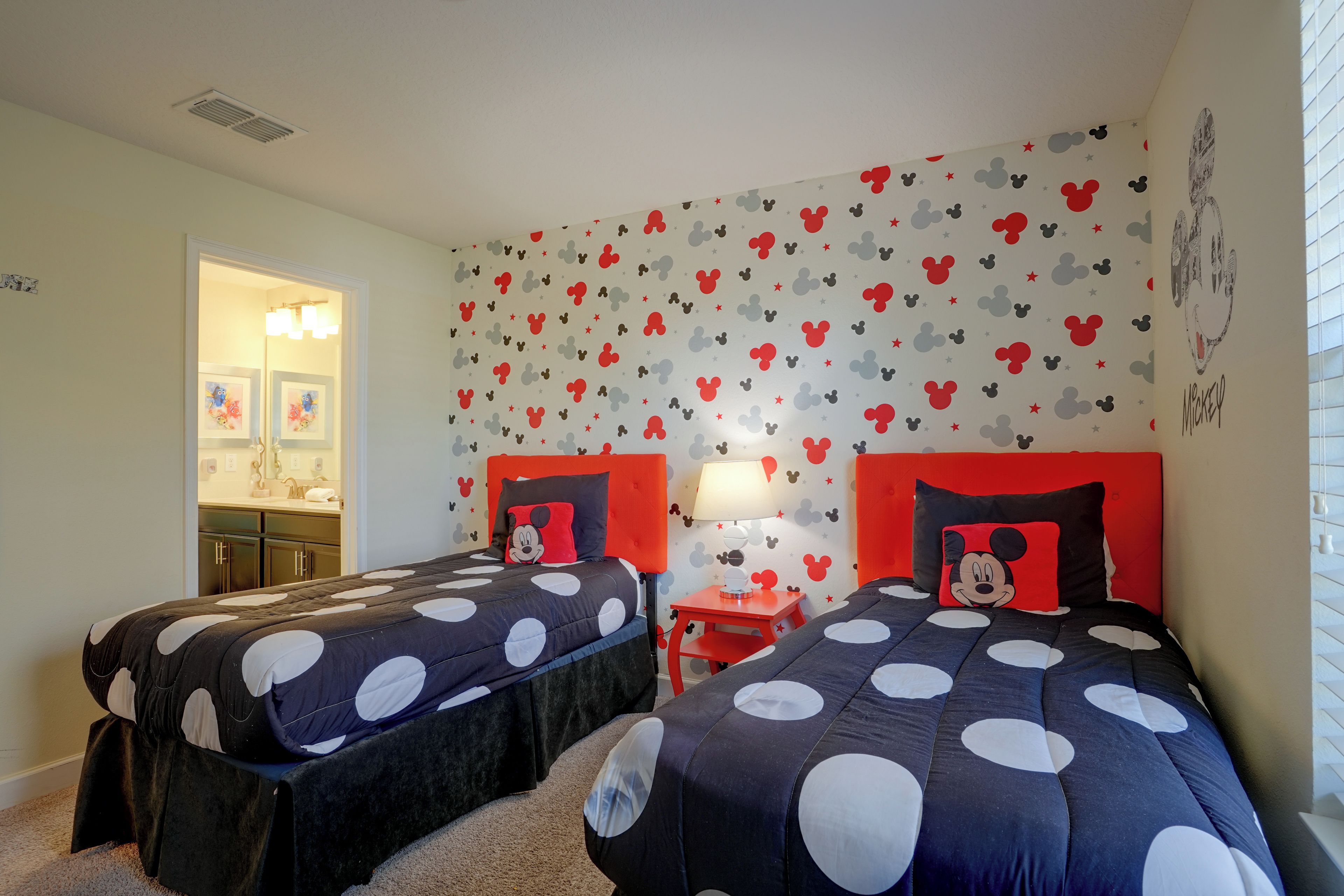 Quarto com duas camas, cada uma com cabeceira temática do Mickey Mouse, mesa de cabeceira com abajur e parede decorada com adesivos do Mickey Mouse e estrelas.