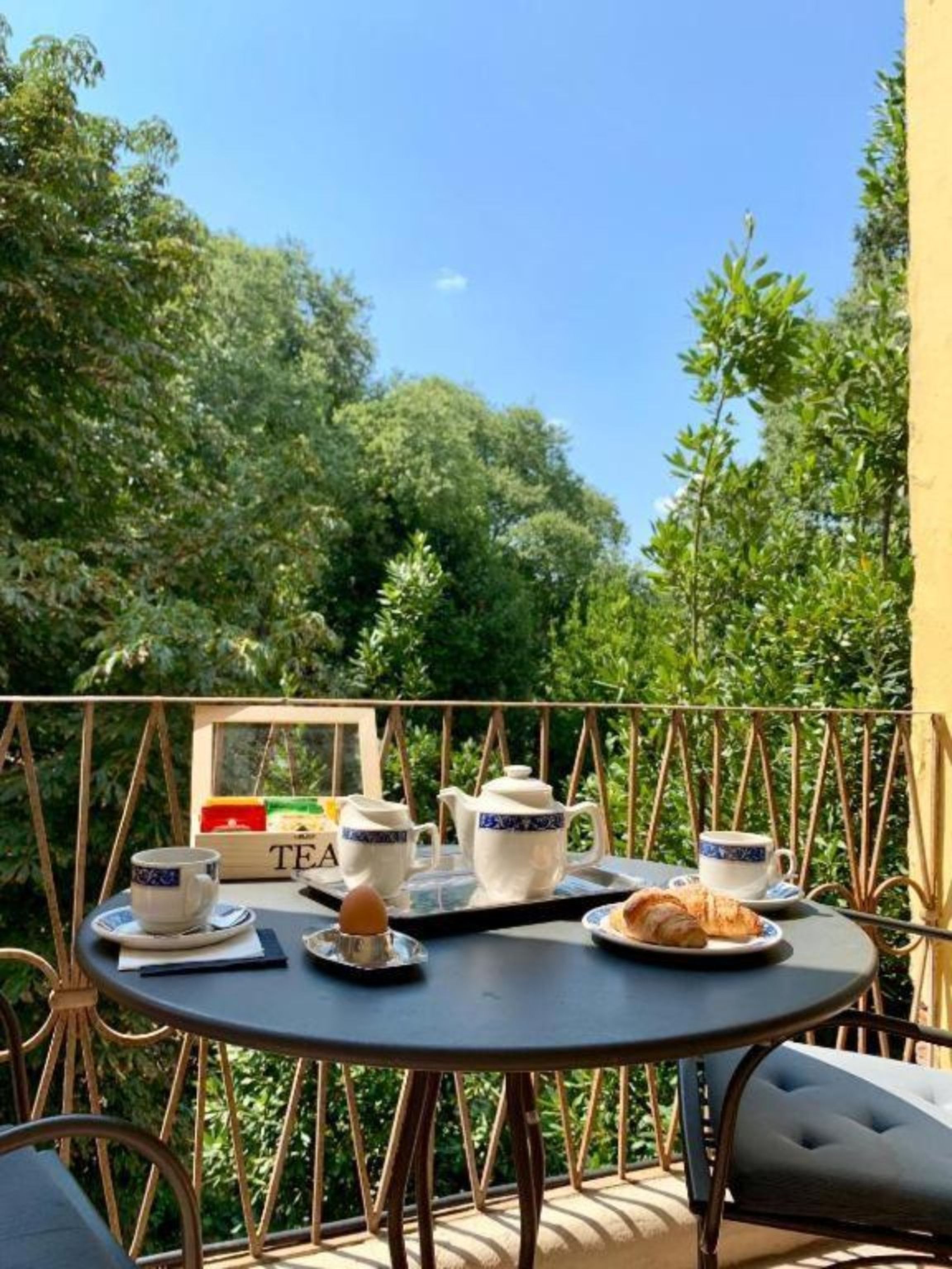 A round table with a tray of tea set, a croissant, and a cup of tea on a balcony with a view of lush greenery.