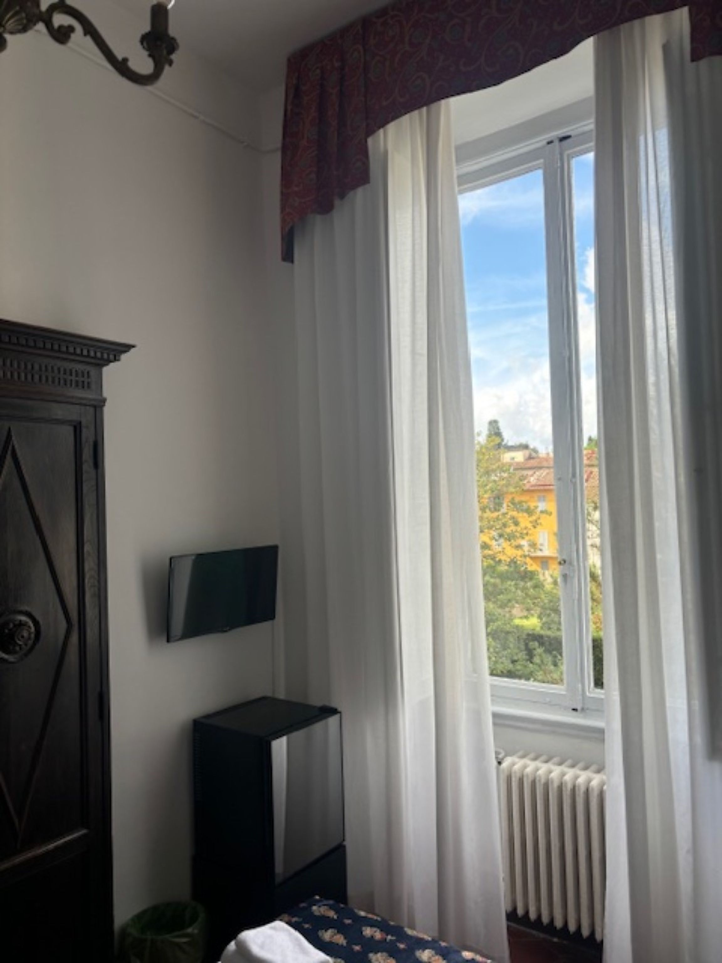 Una habitación con cama, televisor en la pared, radiador y ventana con cortinas.