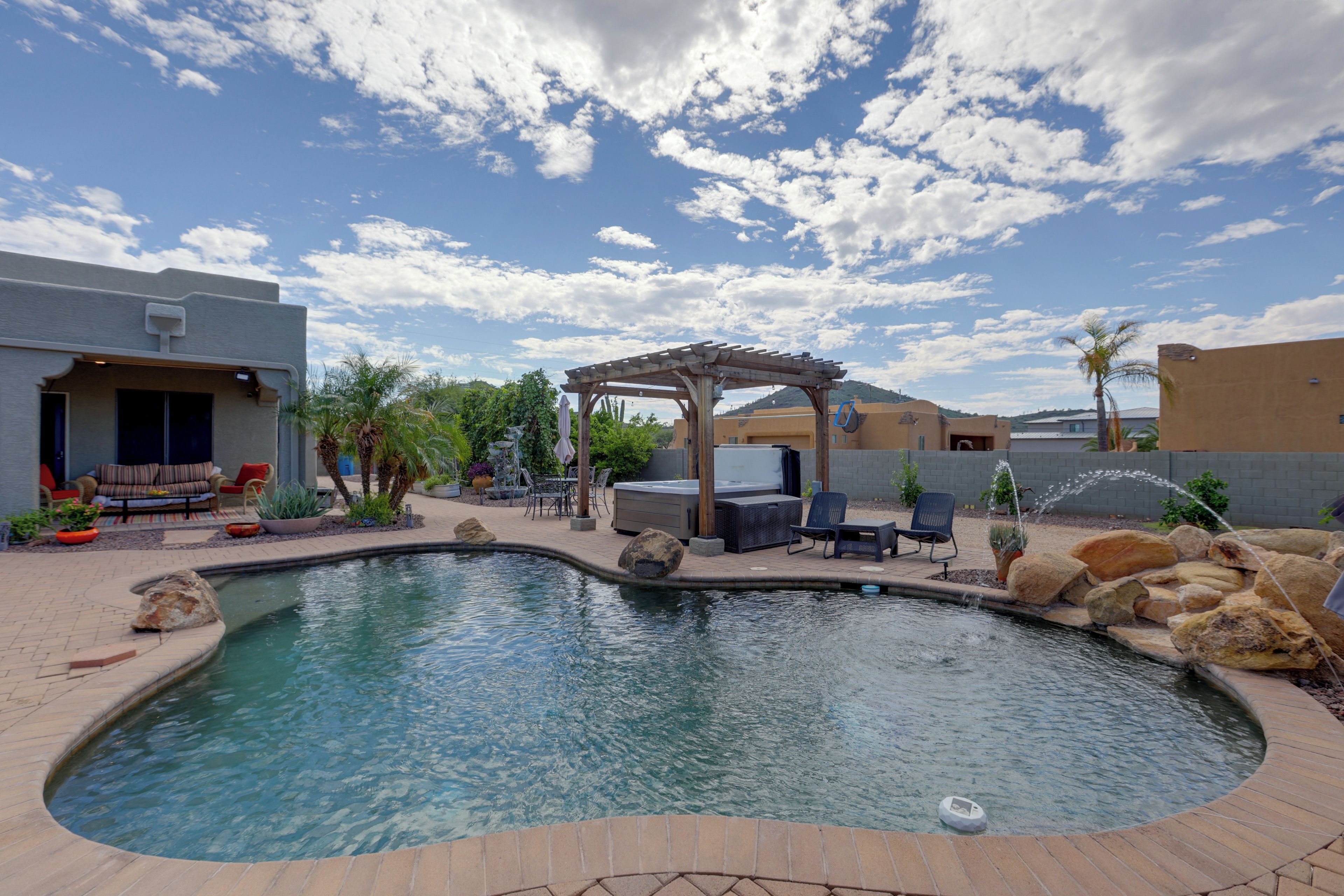 A swimming pool with a hot tub, patio furniture, and a pergola under a partly cloudy sky.