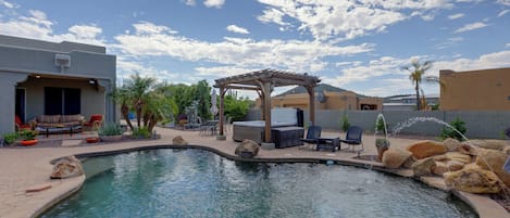 A swimming pool with a hot tub, patio furniture, and a pergola under a partly cloudy sky.