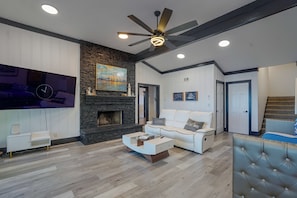 A modern living room with a stone fireplace, white sofas, a flat-screen TV, and a ceiling fan. - Private Dock! Luxury Beachfront Lake Lanier Escape (Buford)