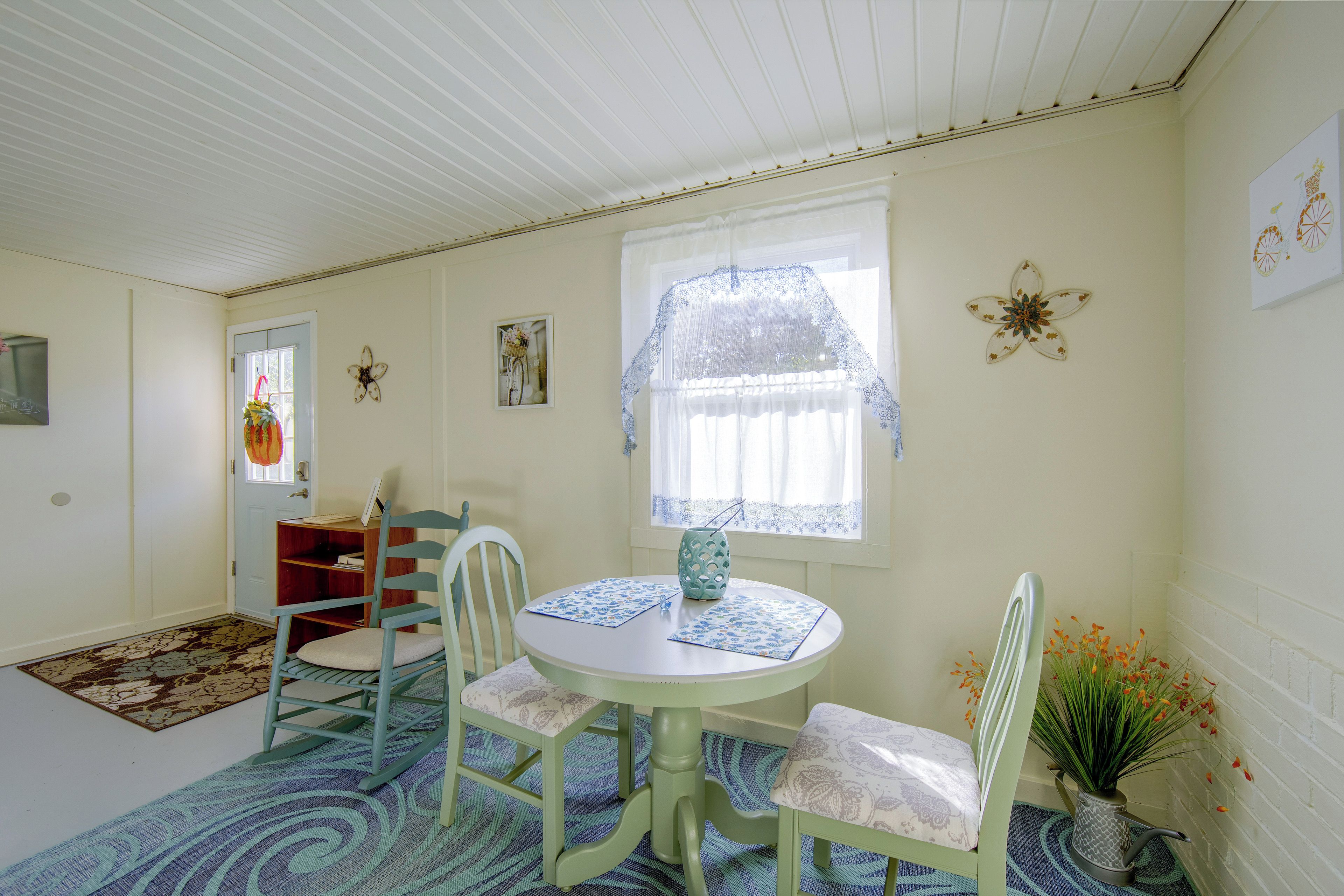 A room with a round table, two chairs, a window with lace curtains, a door, and a wall clock.