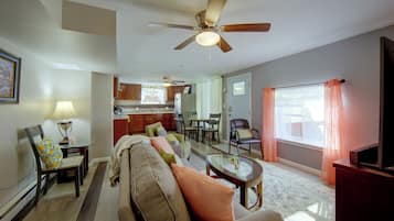 A living room with a sofa, a dining table, a kitchen in the background, and a ceiling fan.