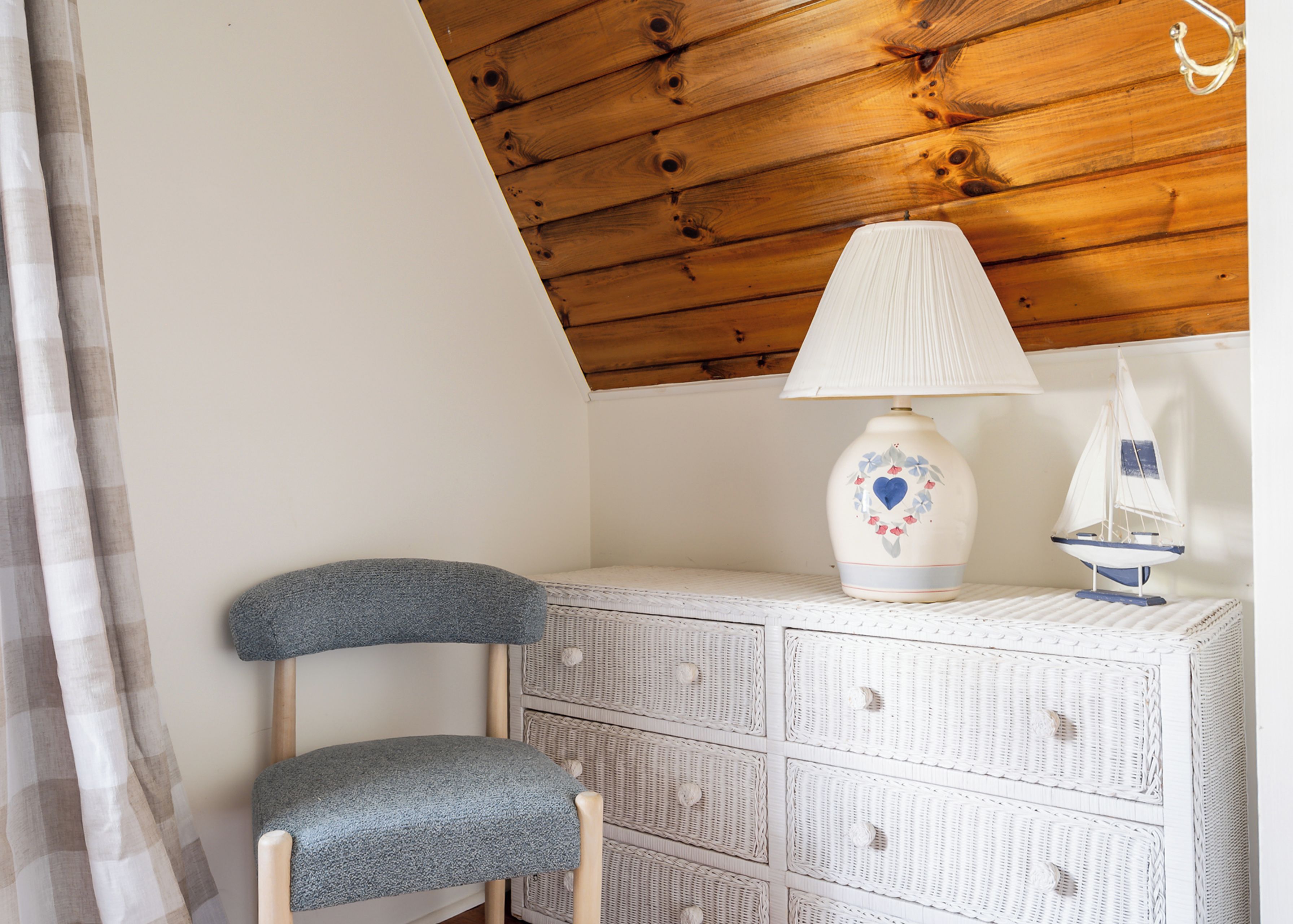 A room with a sloped ceiling, a wicker storage unit, a grey upholstered chair, and a decorative lamp with a sailboat design.