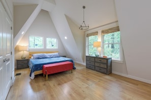 A bedroom with a sloped ceiling, a bed with a red bench, wooden flooring, and two windows with curtains.