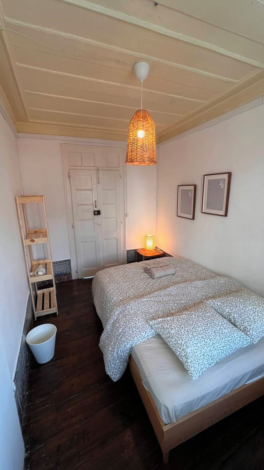 A bedroom with a wooden bed, a hanging light fixture, a small bedside table with a lamp, and a white door.