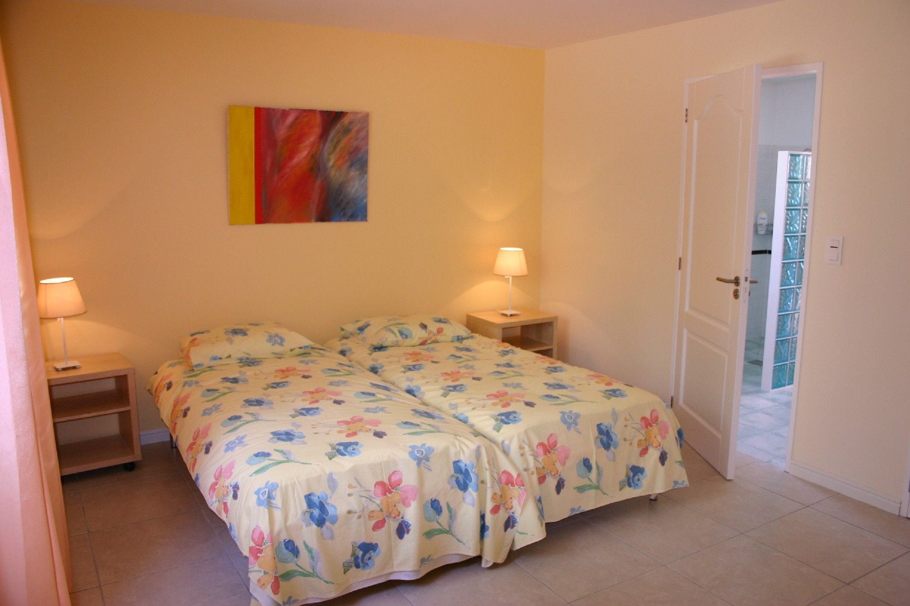 A bedroom with a double bed, two bedside tables, a nightstand, a door to a balcony, and a colorful abstract painting on the wall.