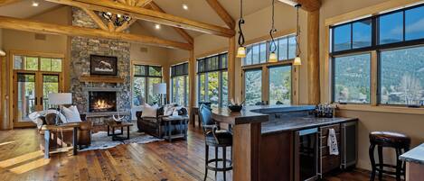A spacious living area with a fireplace, wooden beams, and large windows offering a view of the outdoors.