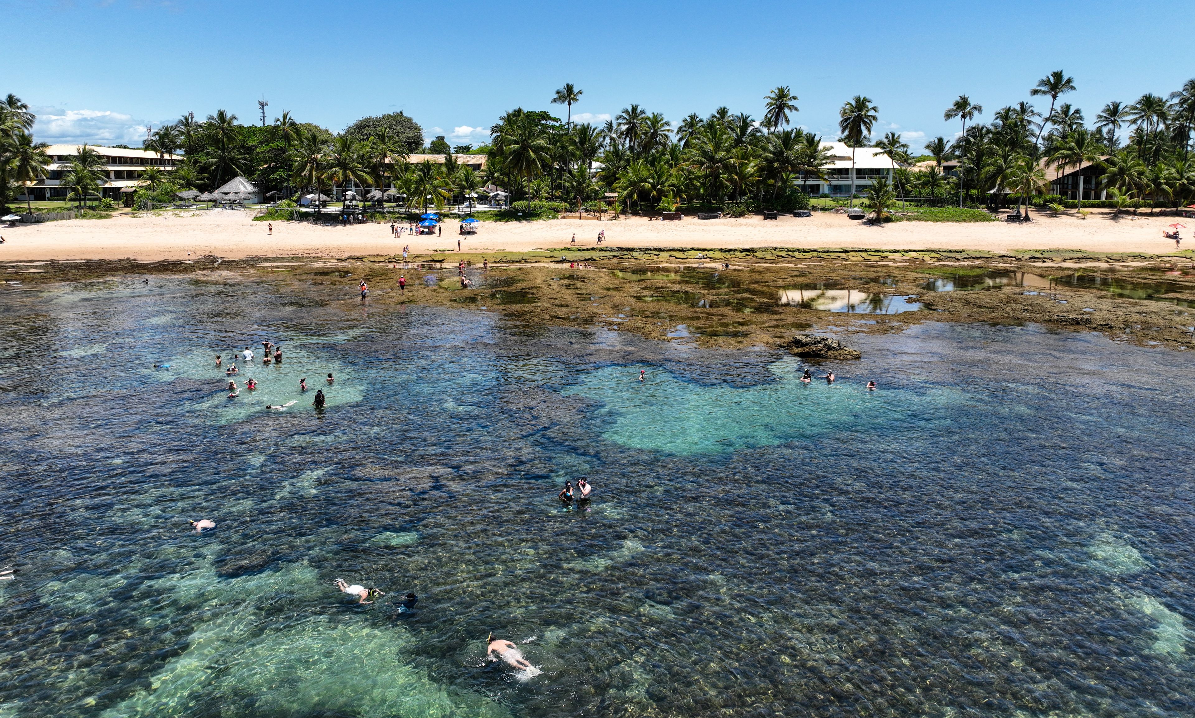 A beach with clear water, palm trees, and a building in the background.