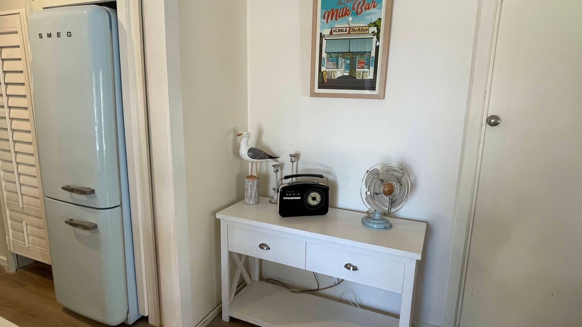 Een keuken in vintage stijl met een witte koelkast, een wit aanrecht met een radio en een ventilator, en een ingelijst beeld aan de muur.