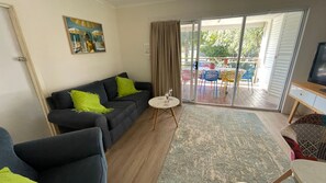 A living room with a sofa, a small round table with two glasses, a television on a stand, and a sliding glass door leading to an outdoor area with a table and chairs. - Super Cool Retro Shack (Cowes)