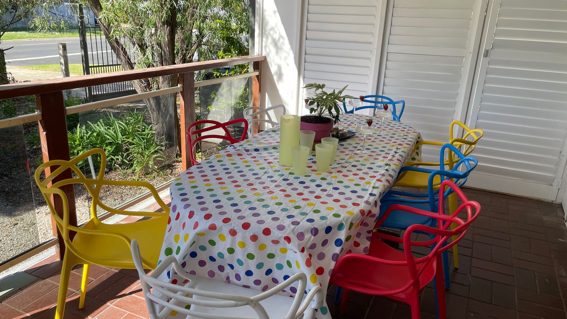 An outdoor patio with a table set for two, surrounded by colorful chairs, a potted plant, and a glass of wine.