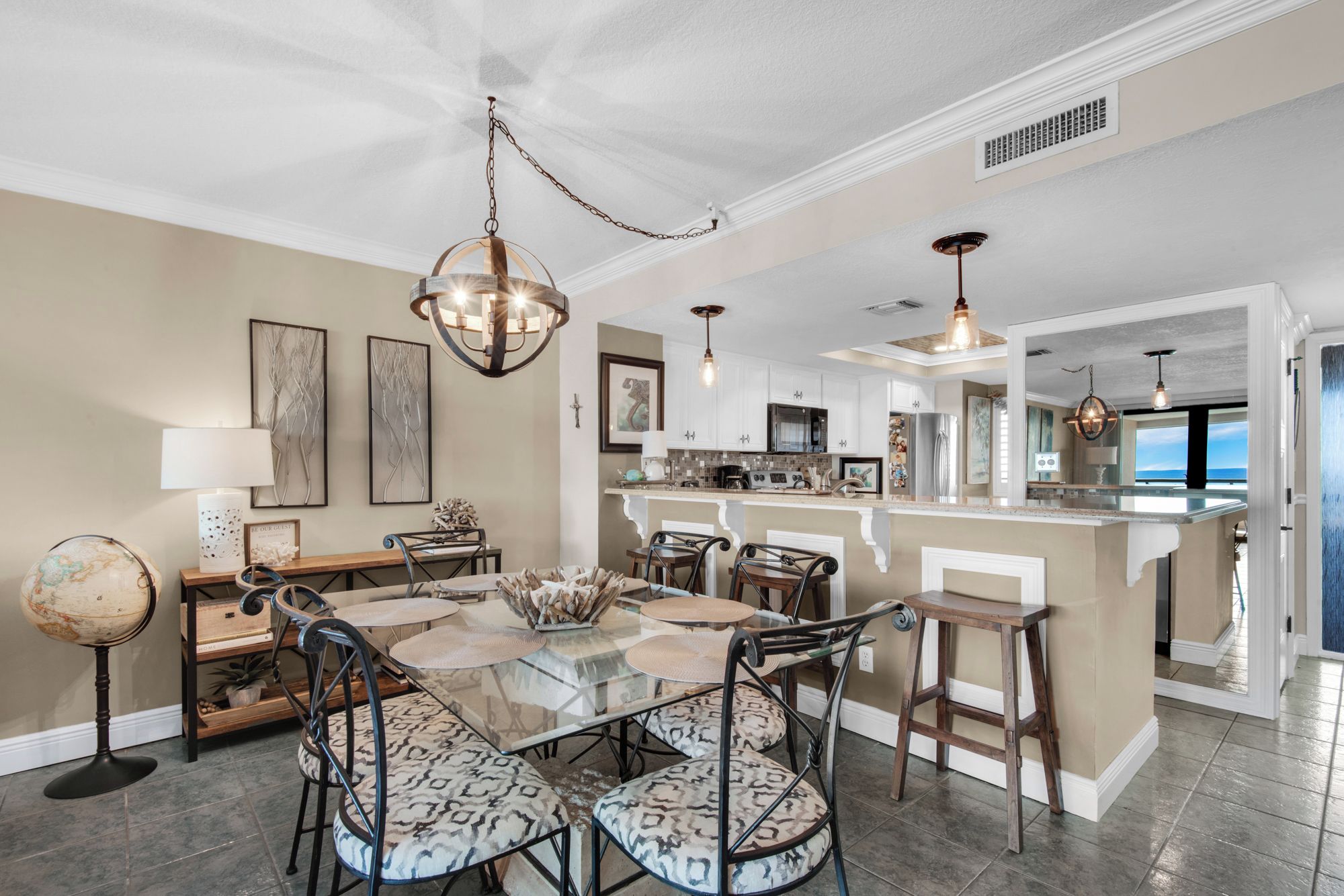 A spacious dining area with a glass table and patterned chairs, a kitchen with white cabinets, and a view of the ocean through a large window.