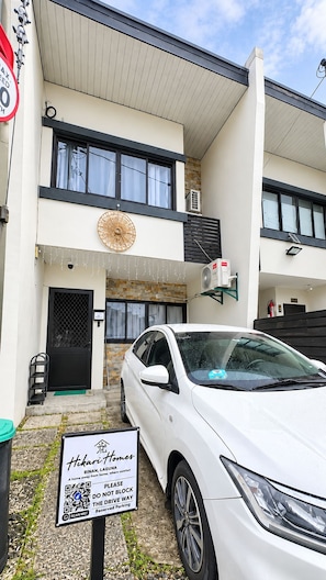 A two-story building with a white car parked in front, a sign for Hikari Homes, and a driveway leading to a black door.