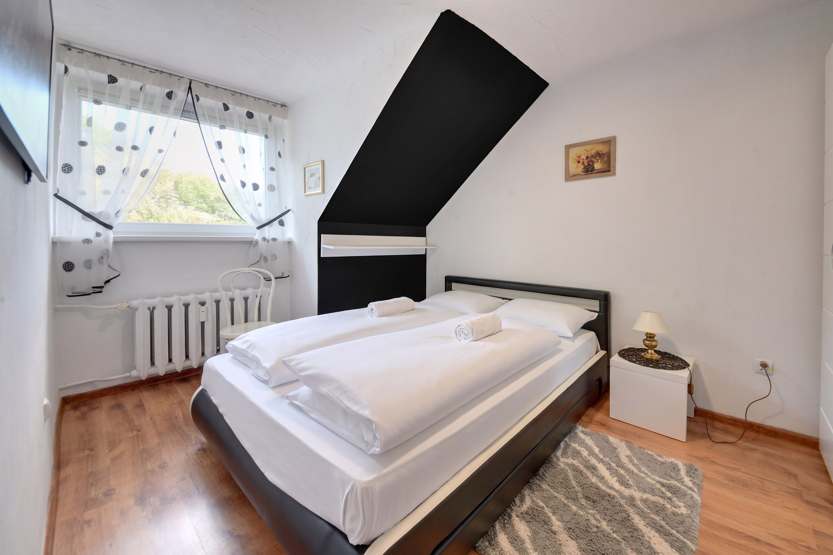 Un dormitorio con cama, radiador, ventana con cortinas, un cuadro en la pared y una mesita de noche con lámpara.