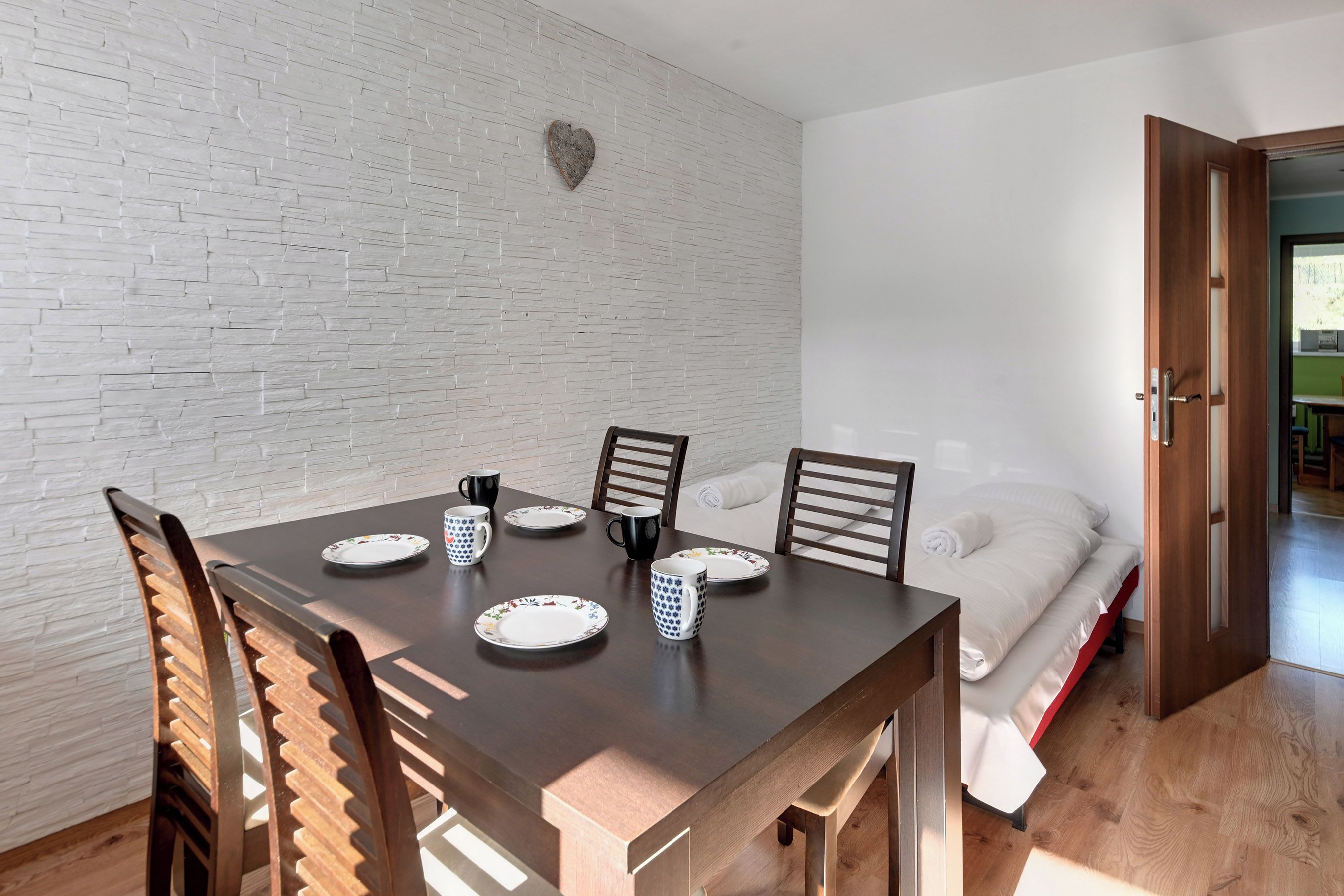 Una taula de menjador amb cadires, una decoració amb forma de cor a la paret i un llit amb edredó blanc a l'habitació propera.