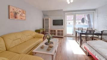 Un saló modern amb un sofà groc, una taula de centre amb un vas, una televisió i una zona de menjador amb una taula i cadires.