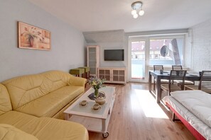 A modern living room with a yellow sofa, a coffee table with a vase, a TV, and a dining area with a table and chairs. - RentPlanet - Apartamenty Wyszyńskiego (Świeradów-Zdrój)