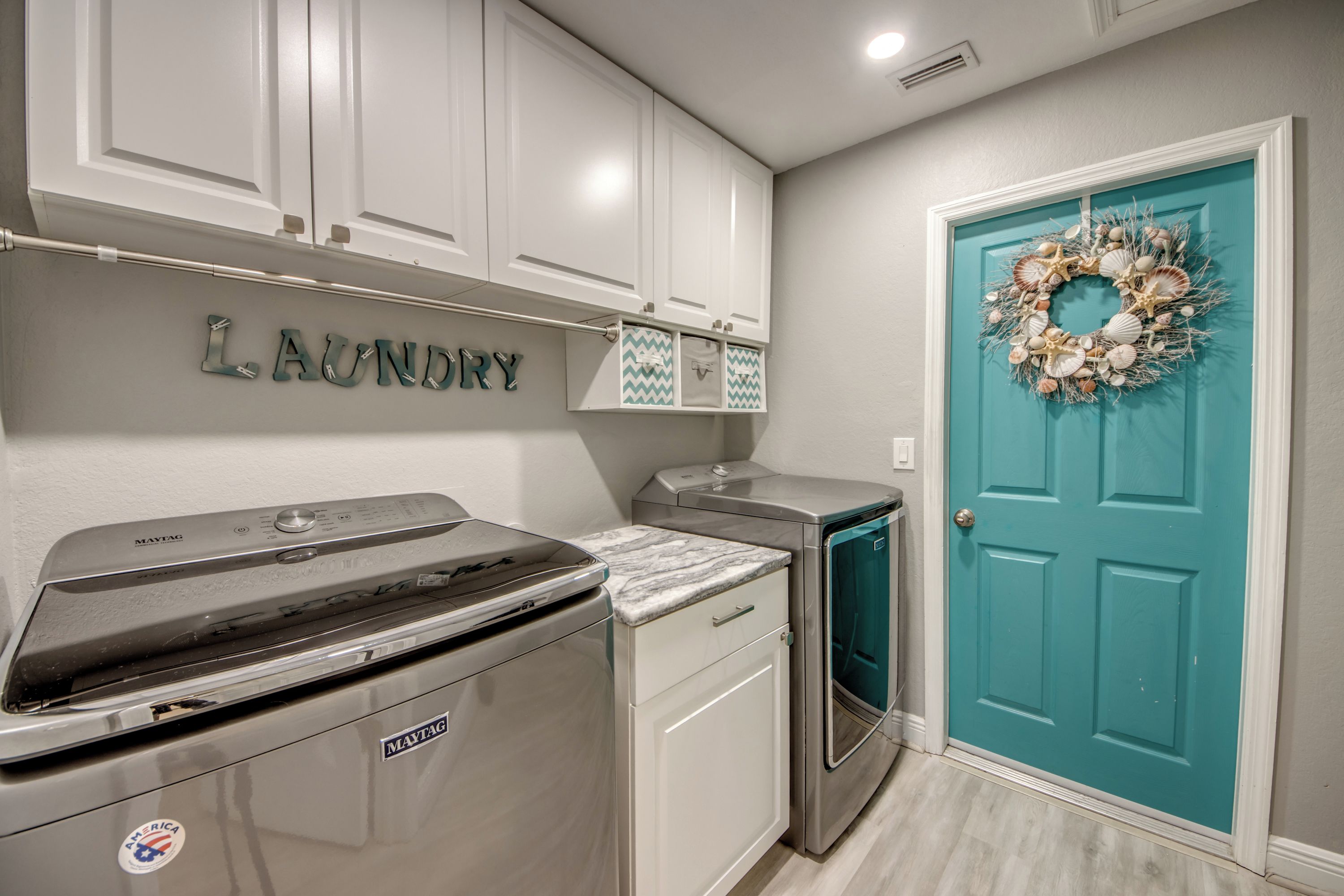 A laundry area with a Maytag washer and dryer, a teal door with a decorative wreath, and a wall-mounted rack with a 'Laundry' sign.