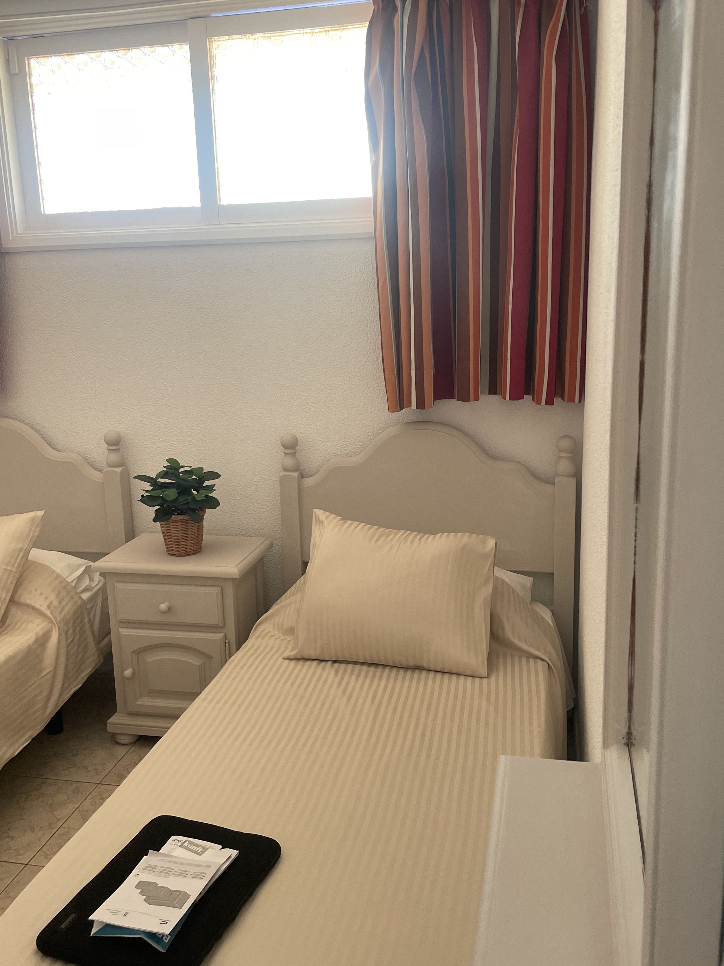 Une chambre avec deux lits, une table de chevet agrémentée d’une plante et une fenêtre avec des rideaux.