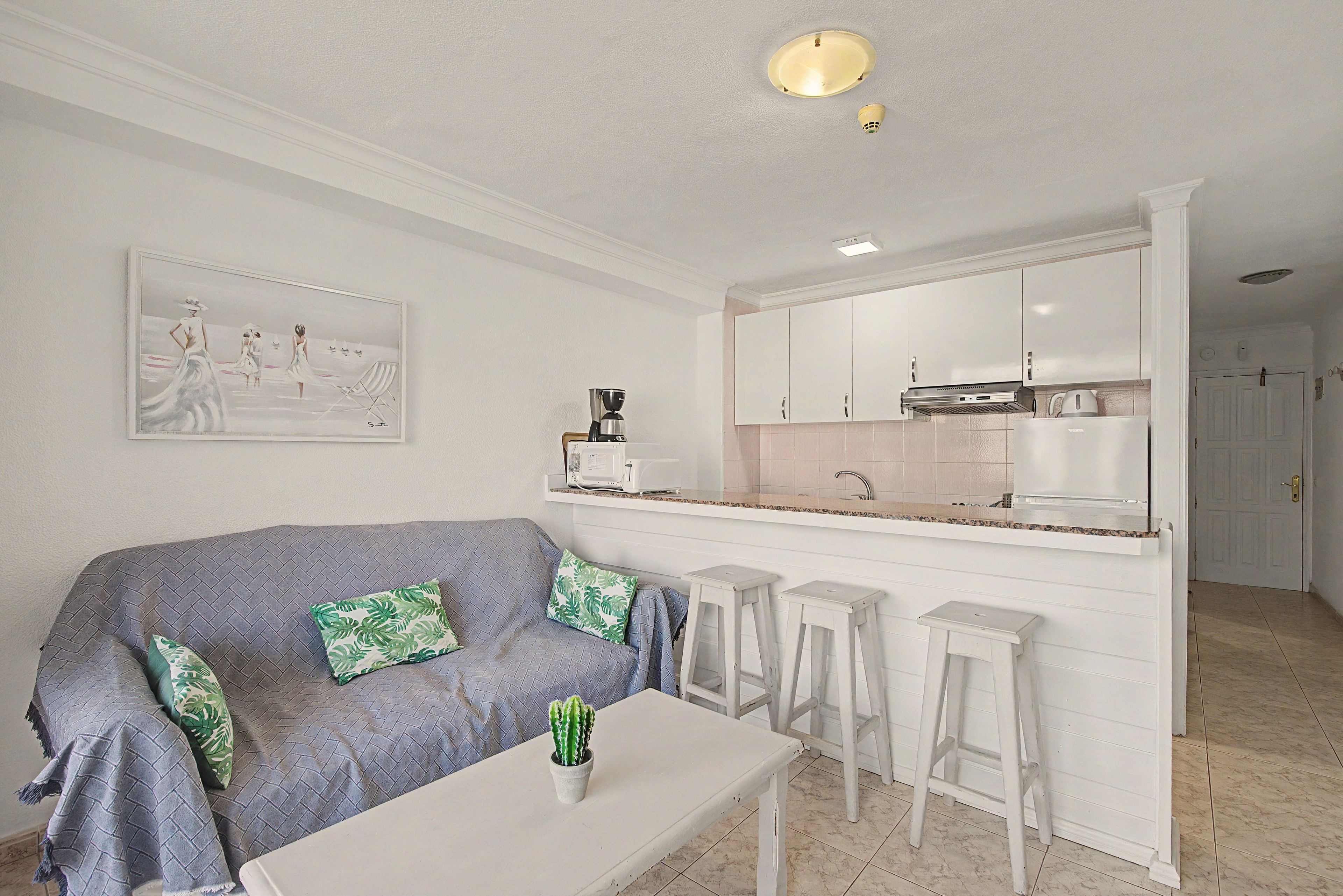 Una cocina moderna con armarios blancos, una cafetera y un área de comedor con sofá gris y taburetes blancos.