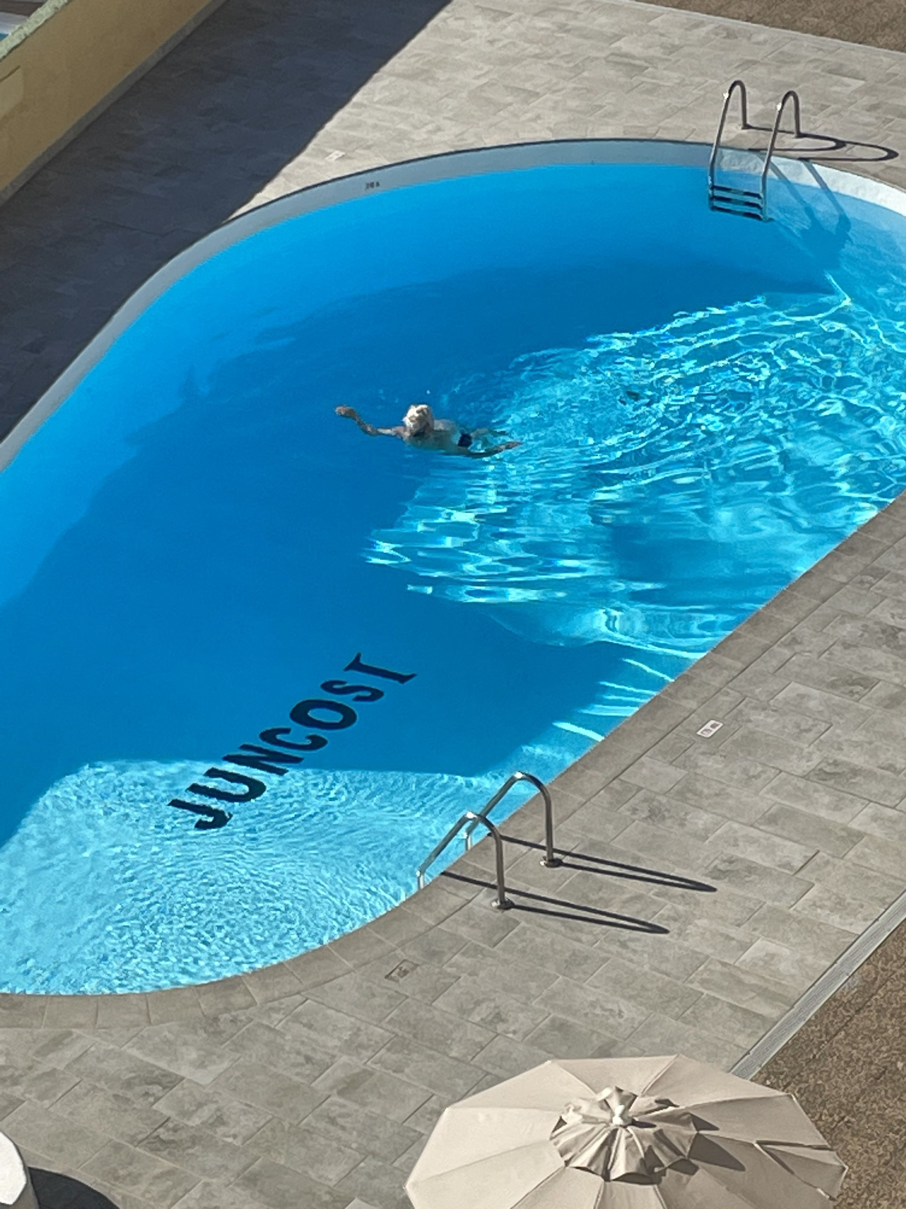 Piscina com uma pessoa nadando, um guarda-sol à beira da piscina e a palavra 'JUNCOAST' na borda da piscina.