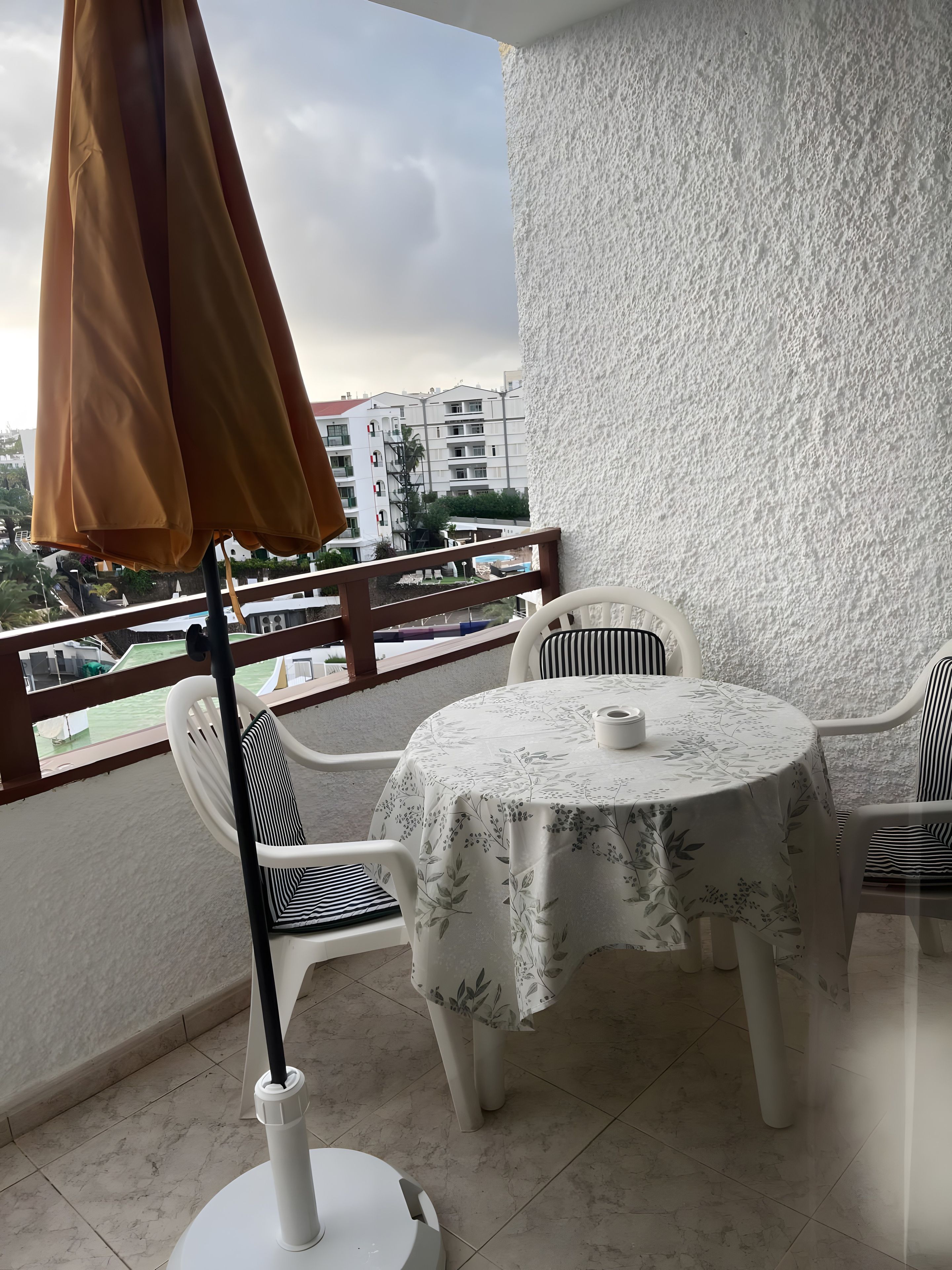 Un balcon avec une table et des chaises, un mur blanc à la texture intéressante et une vue sur des bâtiments et de la verdure.