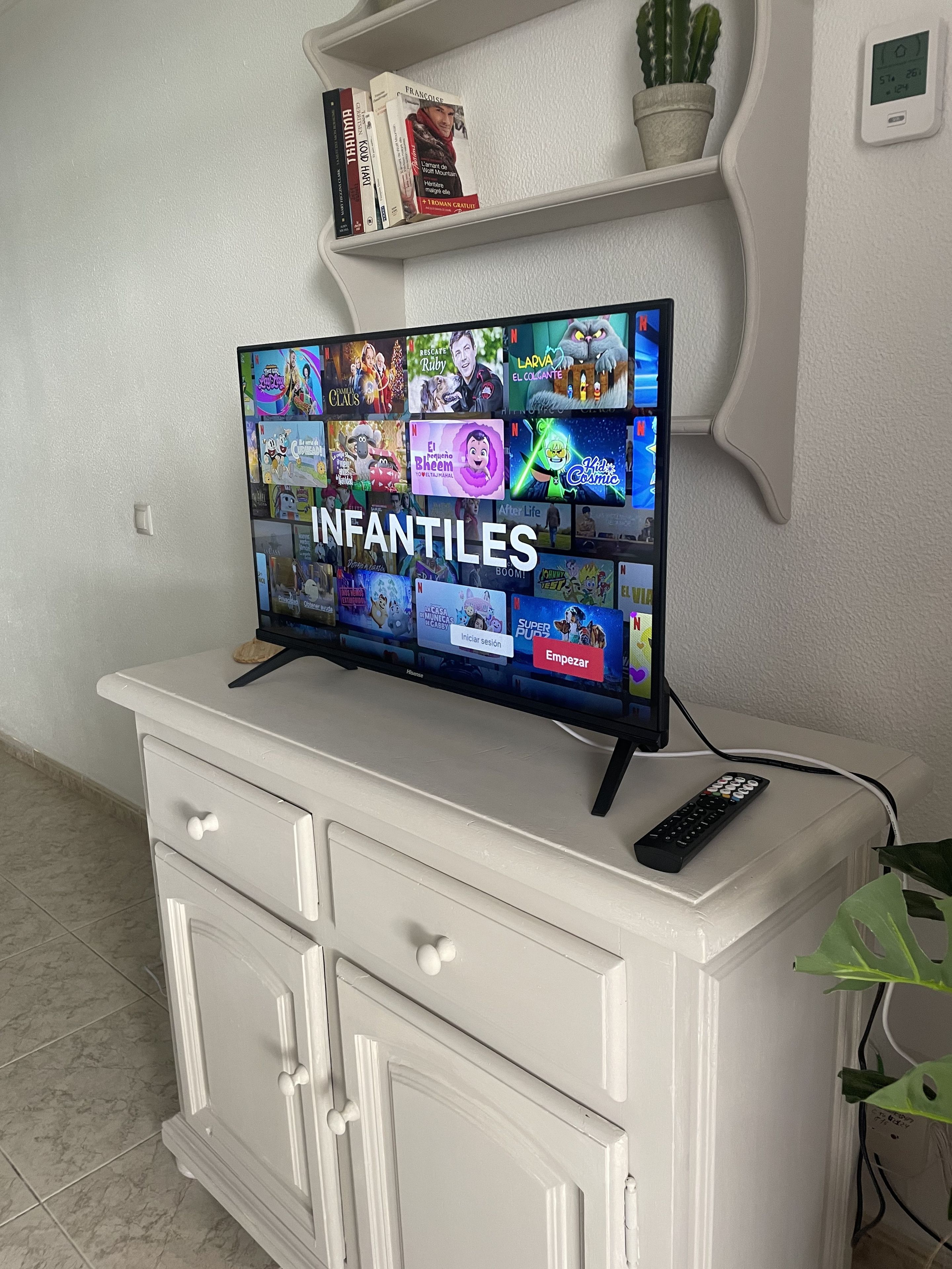 Een televisie op een wit kastje, met een afstandsbediening, omringd door boeken en een potplant.