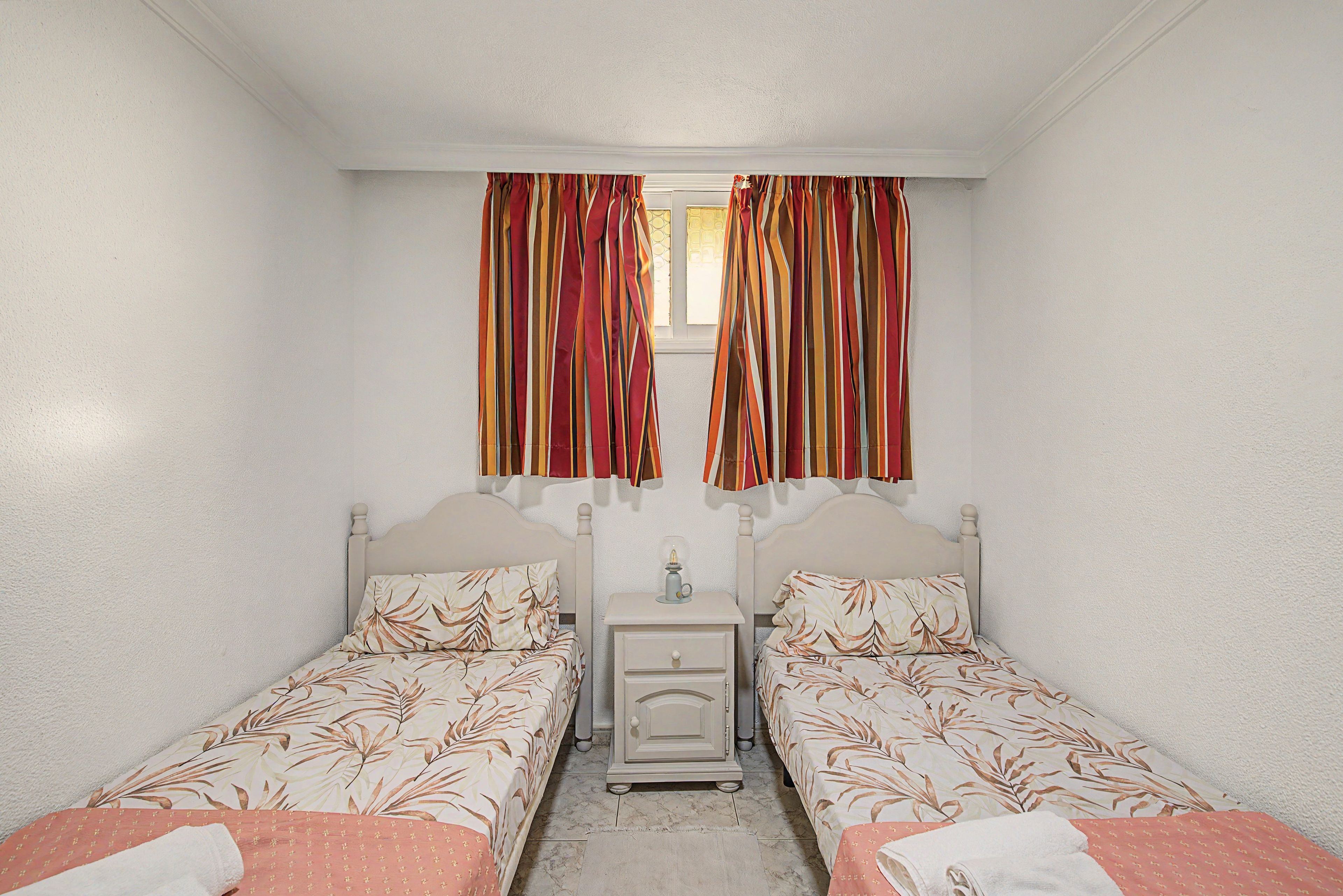Una habitación con dos camas individuales, una mesita de noche, una pequeña mesa y una ventana con cortinas.