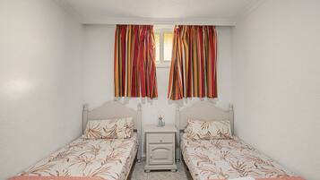 Una habitación con dos camas individuales, una mesita de noche, una pequeña mesa y una ventana con cortinas.