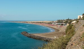 A coastal beach scene with a rocky breakwater, sandy beach, and a town in the background. - 'Villasyaiza ' los Juncos I' (San Bartolomé de Tirajana)