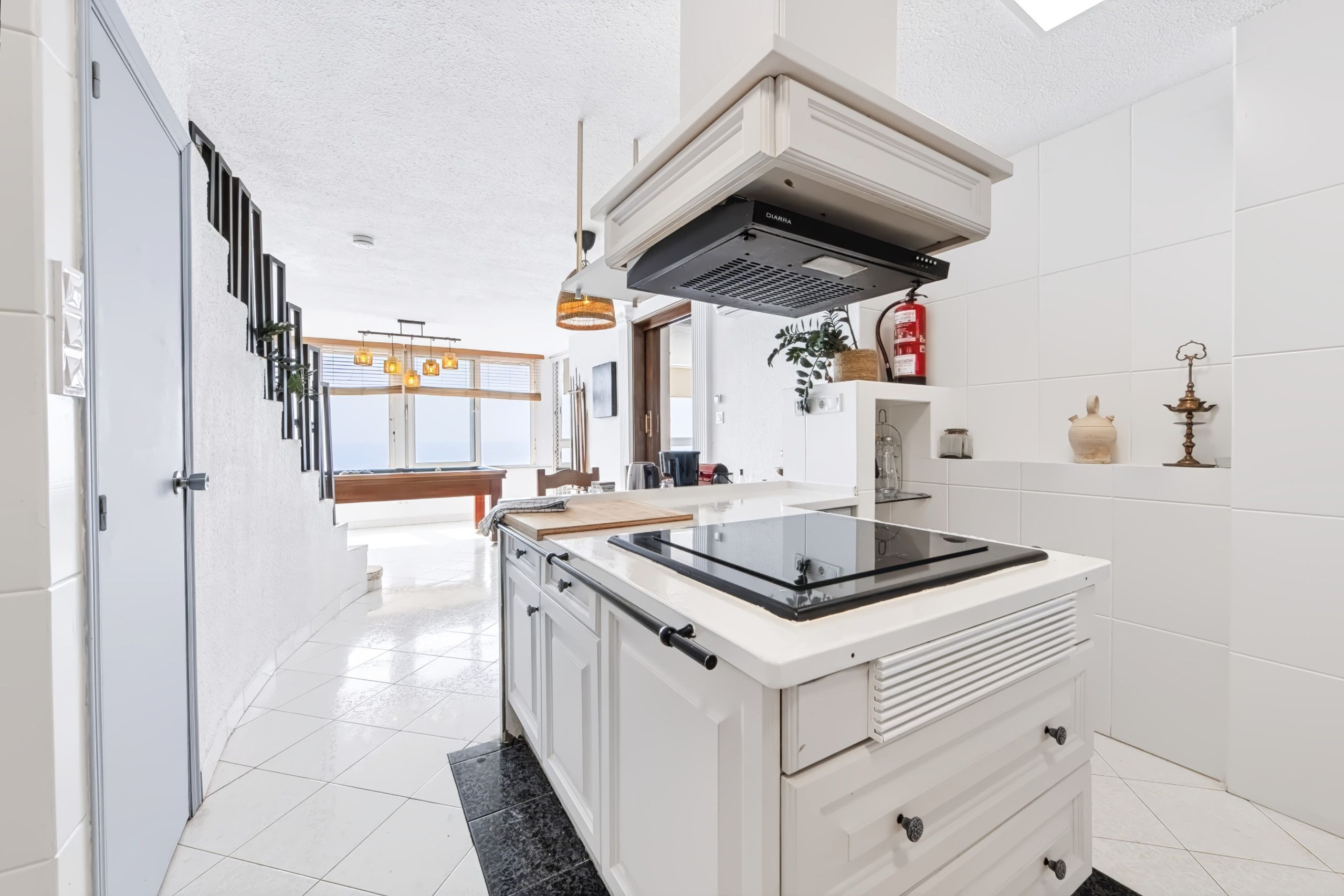 Una cocina moderna con armarios blancos, una placa de cocina negra y un horno empotrado. Hay una ventana que proporciona luz natural y vistas al exterior.