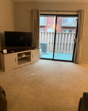 Ein Wohnzimmer mit einem Flachbildfernseher auf einem Schrank, einer Schiebetür zum Balkon und einem beigen Teppich.