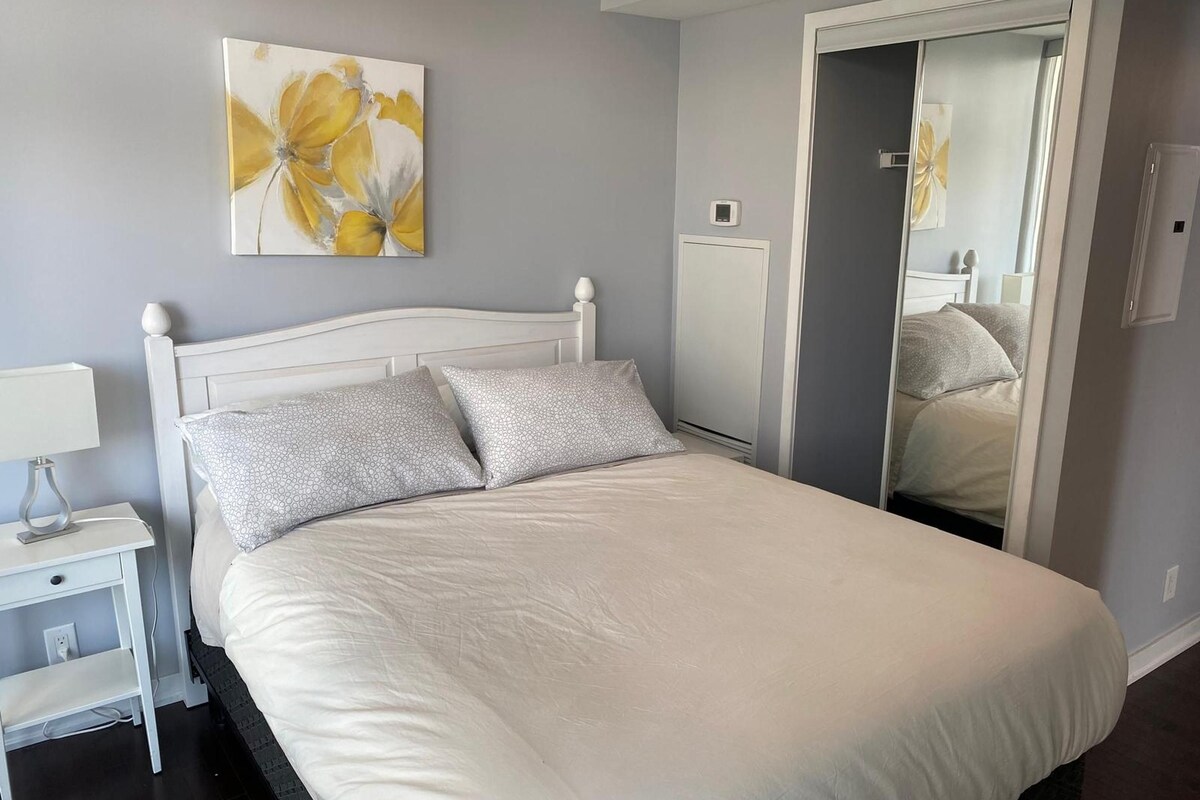 A neatly made bed with white bedding and two pillows, a bedside table with a lamp, and a large mirror reflecting part of the room.