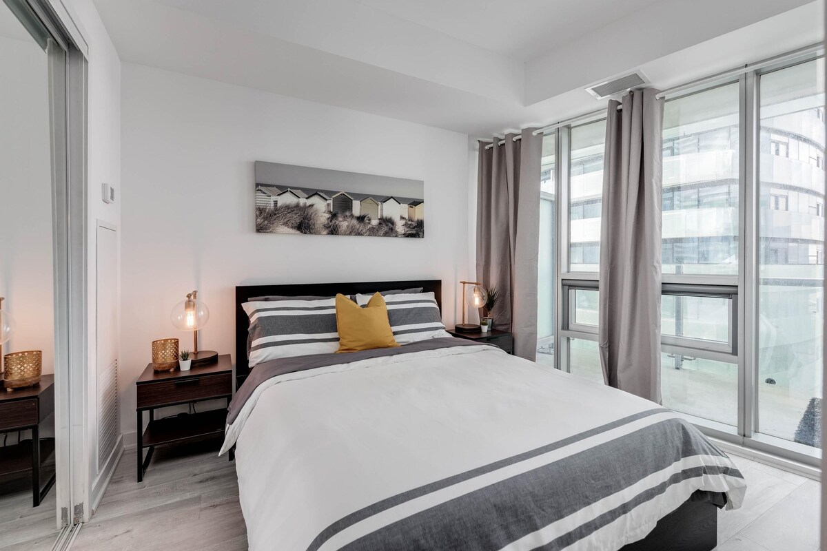 A modern bedroom with a large bed, a nightstand with a lamp, a wall-mounted artwork, and a view through large windows.