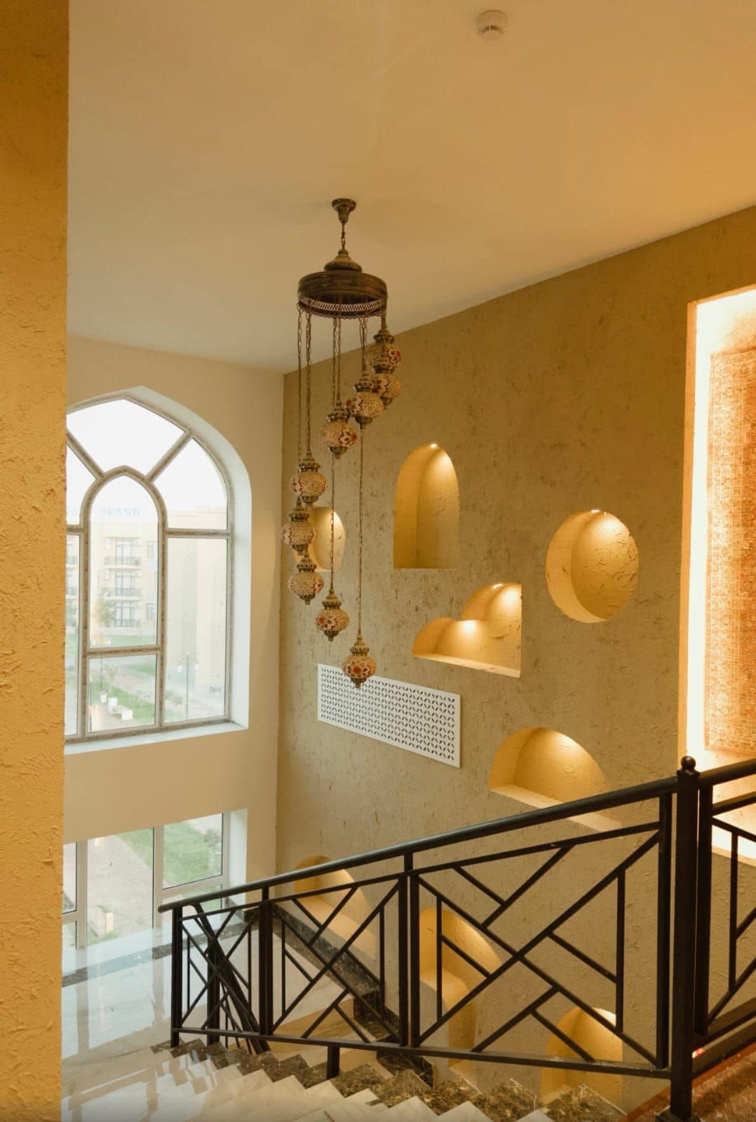 Una escalera con un diseño geométrico en la barandilla, un candelabro decorativo colgante y vistas a través de un amplio ventanal.