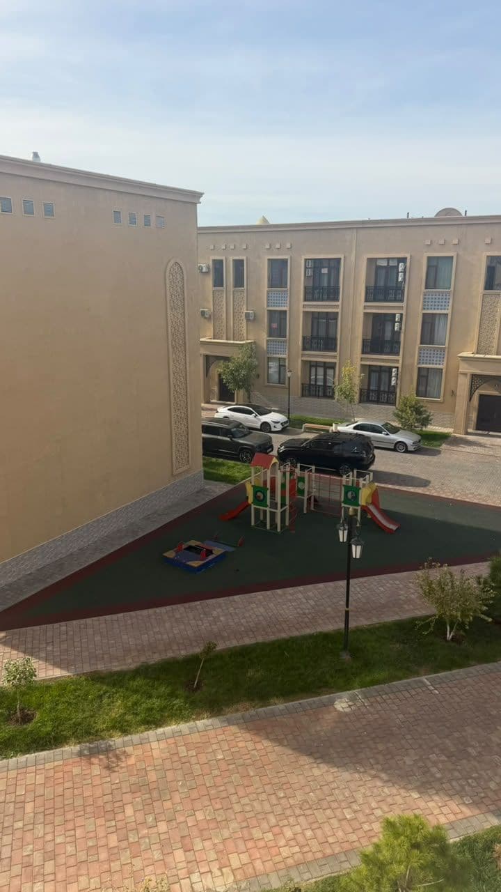 Un parque infantil con un tobogán y estructuras de escalada, rodeado de una zona pavimentada y un edificio con amplias ventanas.