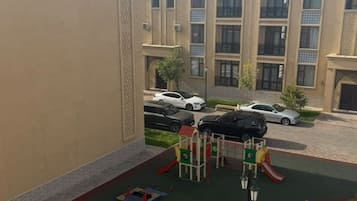 Un parque infantil con un tobogán y estructuras de escalada, rodeado de una zona pavimentada y un edificio con amplias ventanas.