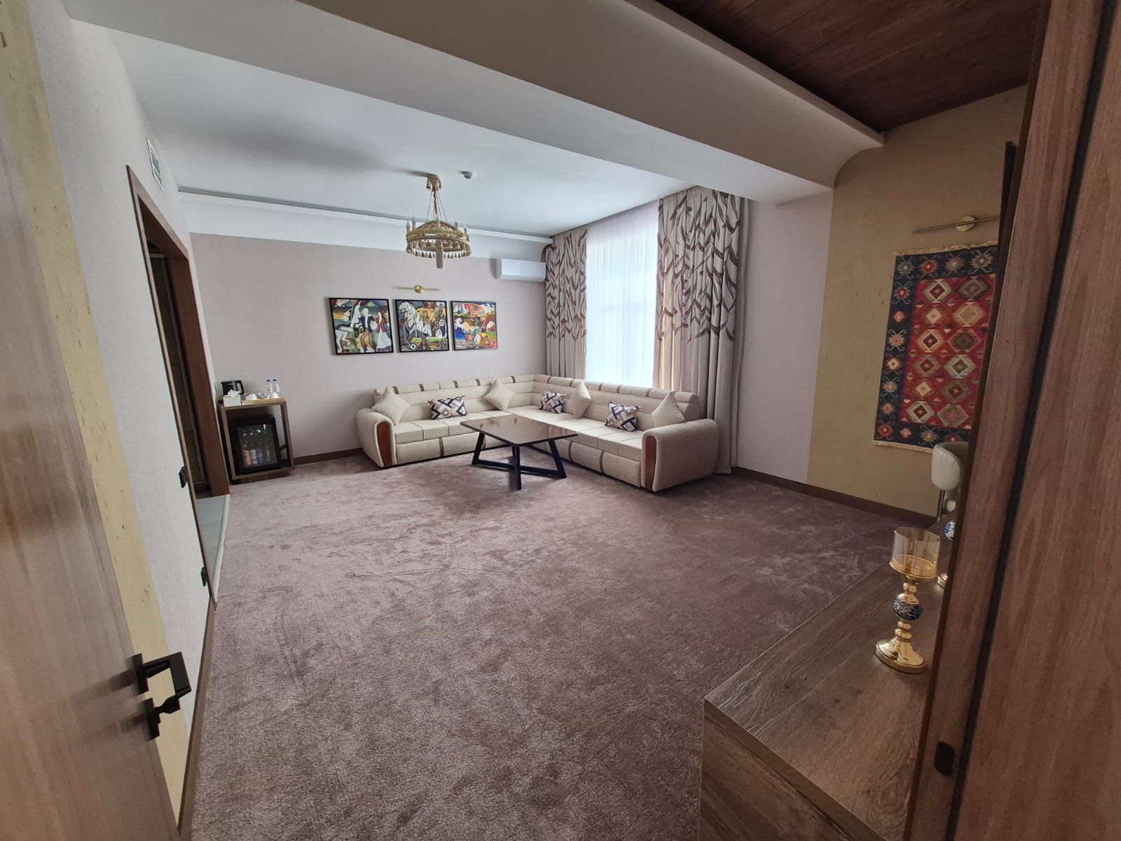 Uma sala de estar com sofá seccional, mesa de centro e lustre. Há três quadros na parede e um tapete estampado.