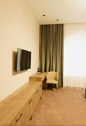 A room with a flat-screen TV mounted on the wall, a wooden desk with drawers, a chair, and a large window with curtains.