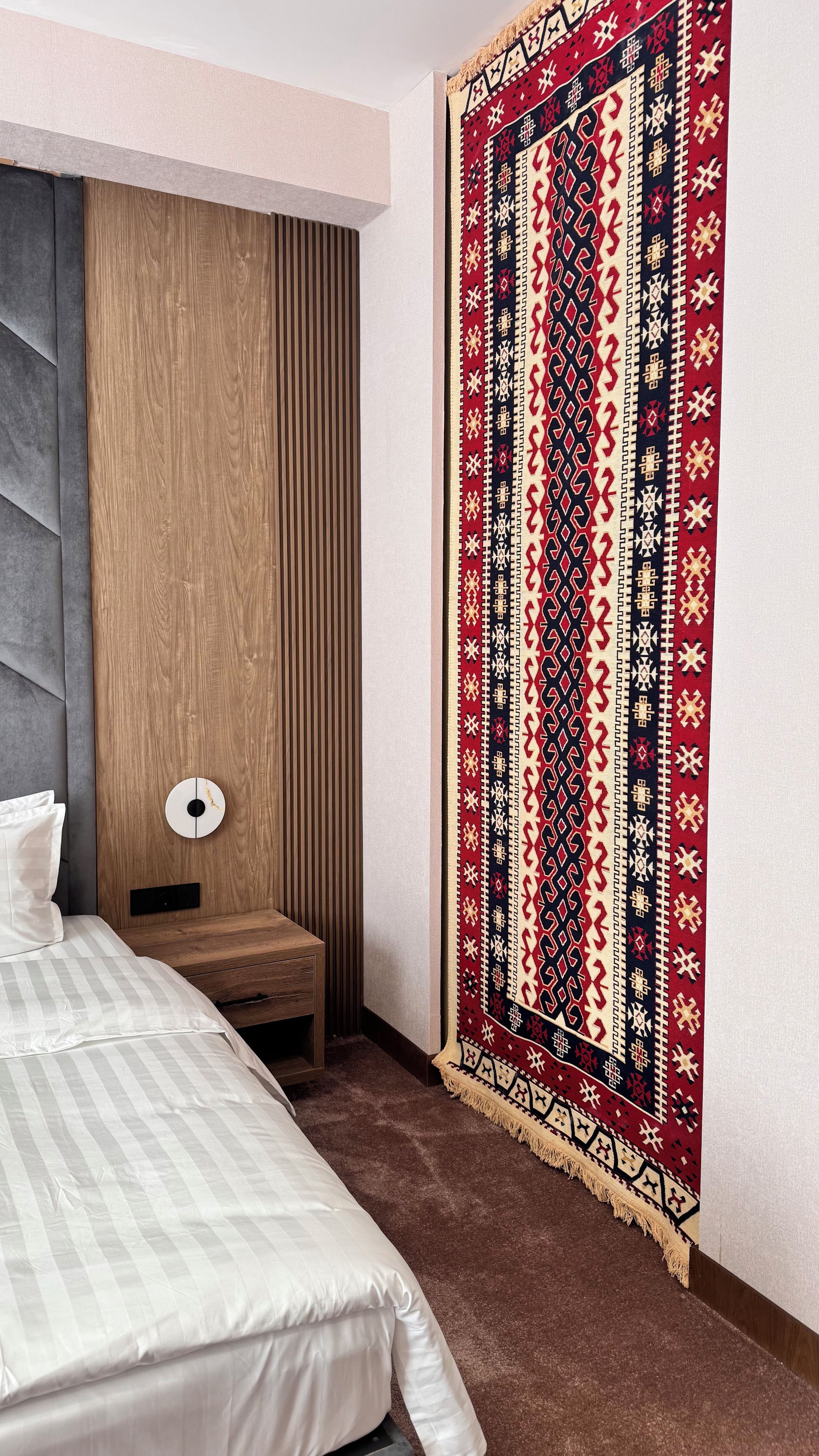 Un dormitorio con una cama, cabecera de madera, mesita de noche, lámpara y un tapiz decorativo en la pared.