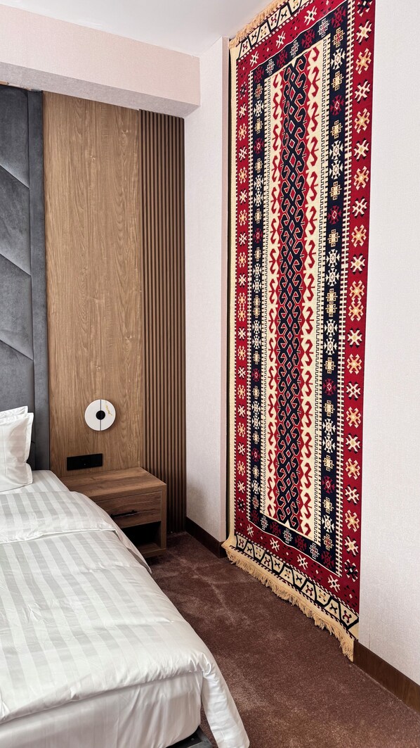 A bedroom with a bed, a wooden headboard, a bedside table, a lamp, and a patterned wall hanging.