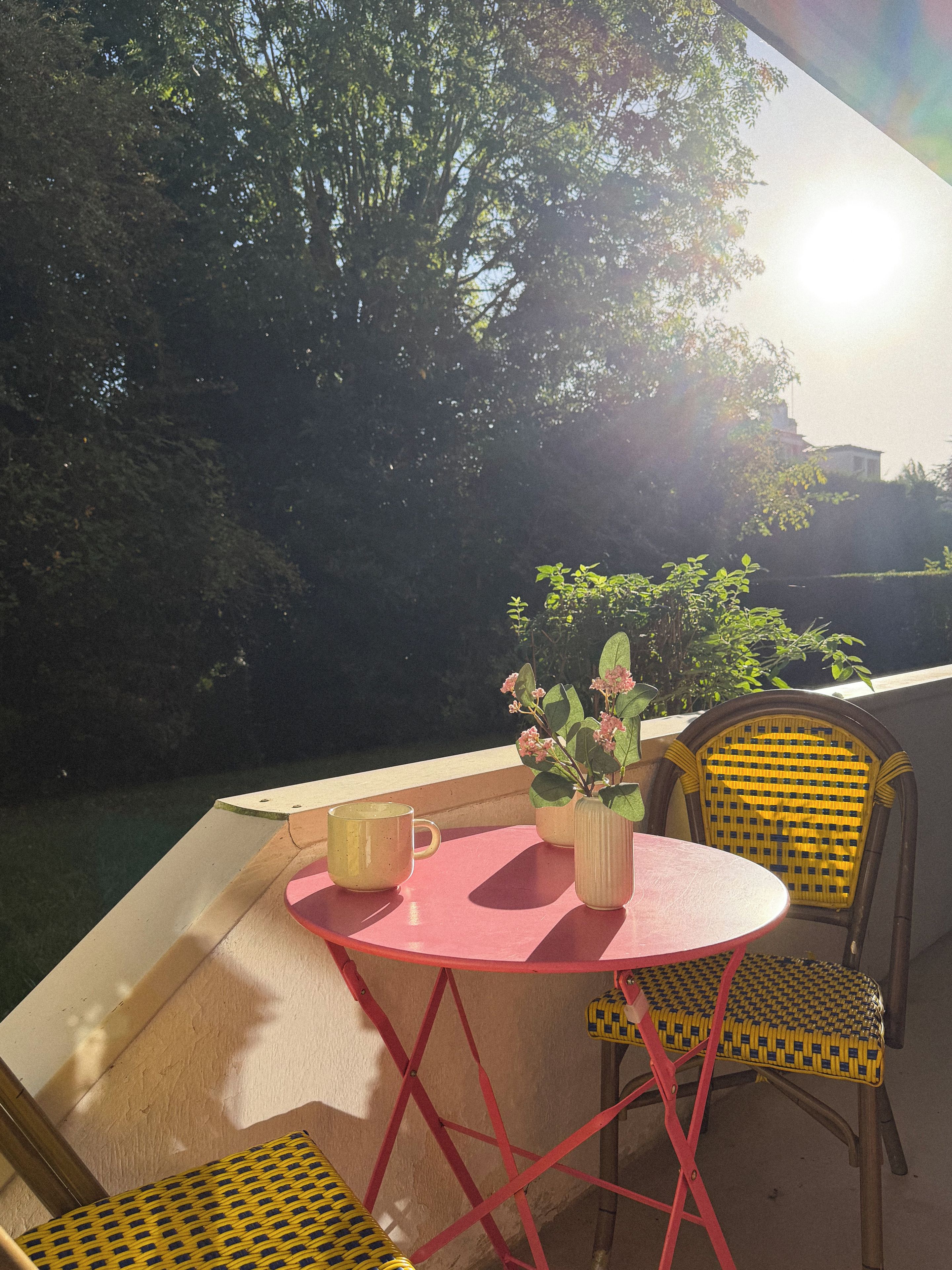 A balcony with a table and chairs, a potted plant, and a view of trees and a building in the background.