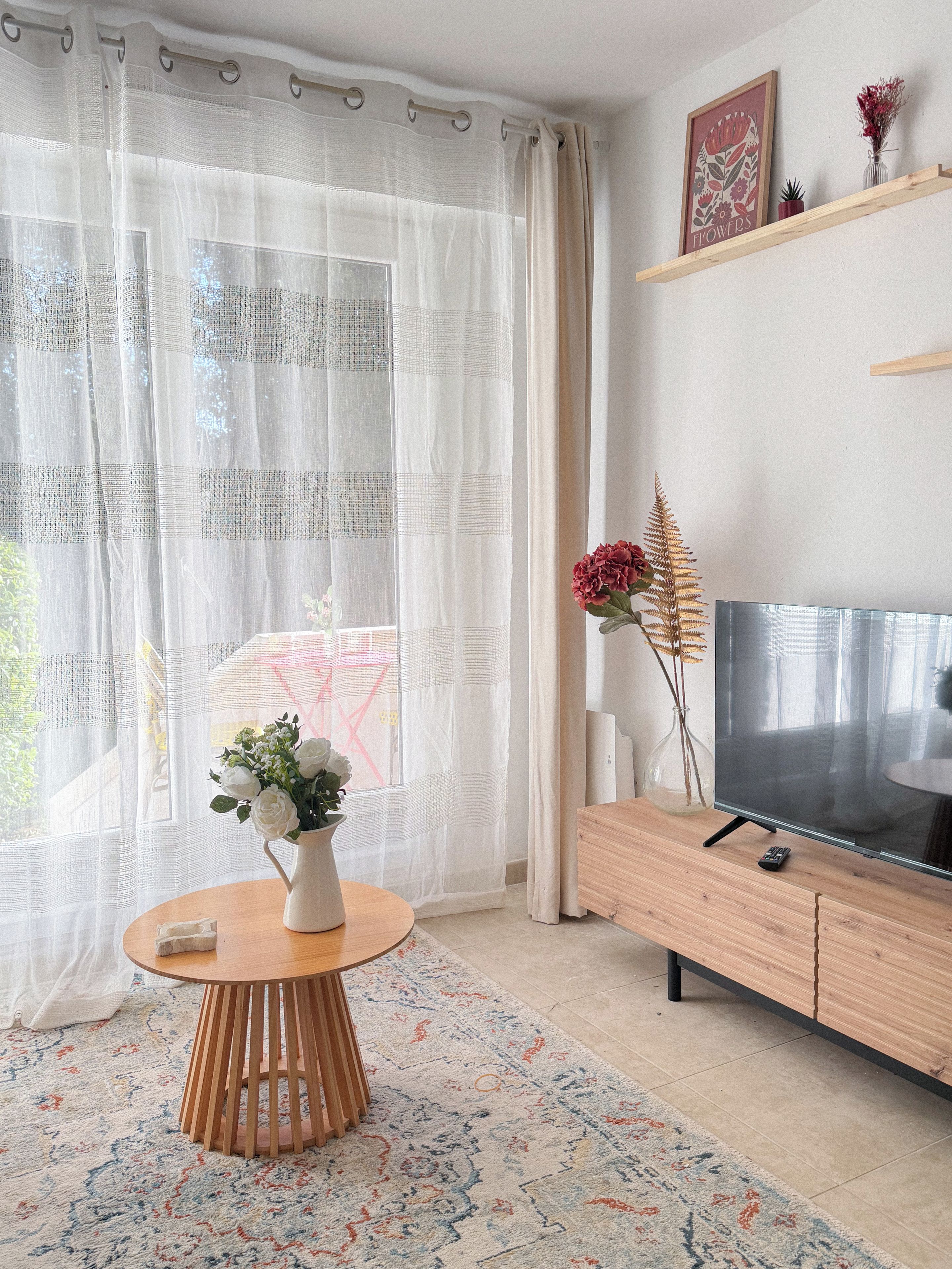 Un salon avec une table ronde en bois, un téléviseur posé sur un meuble en bois et une grande fenêtre ornée de rideaux légers.